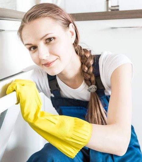 Smiling woman in beige cleaning uniform with yellow gloves holding a spray bottle and blue cloth in a bright living room.