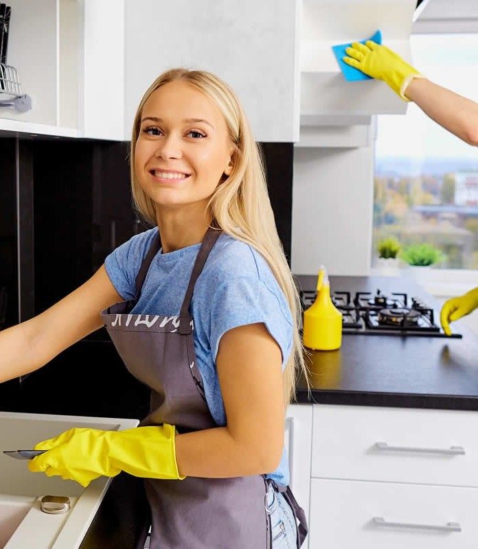 Smiling woman in a beige cleaning uniform wearing yellow gloves and holding a feather duster in a bright living room.