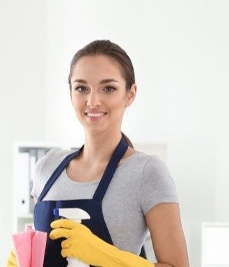 Smiling woman in beige uniform and yellow gloves holding a brush and a gray bucket in a bathroom.