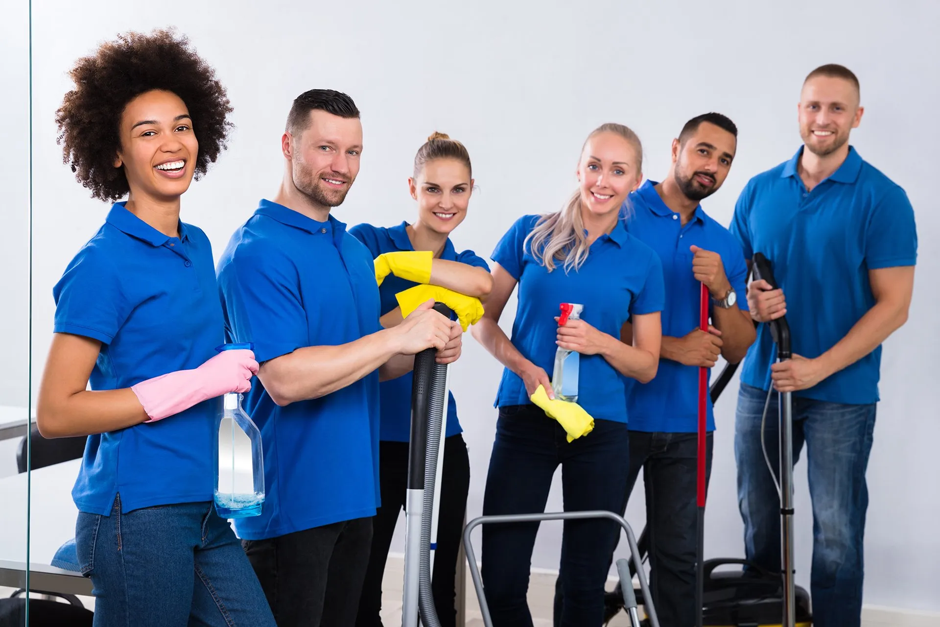 Group of six smiling cleaning staff in beige uniforms holding cleaning supplies in a bright living room.
